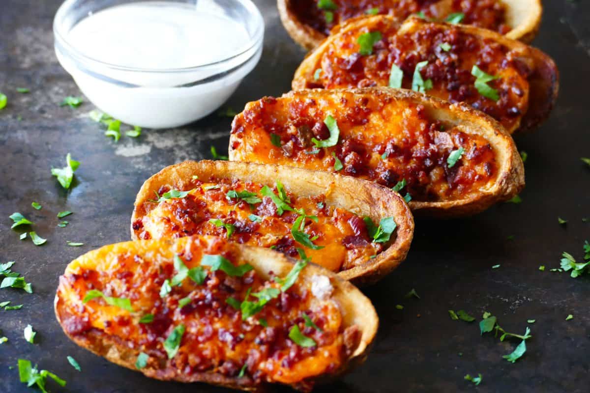 5 potato skins in a row with melted cheese and bacon and garnished with parsley with a glass bowl of white sauce.