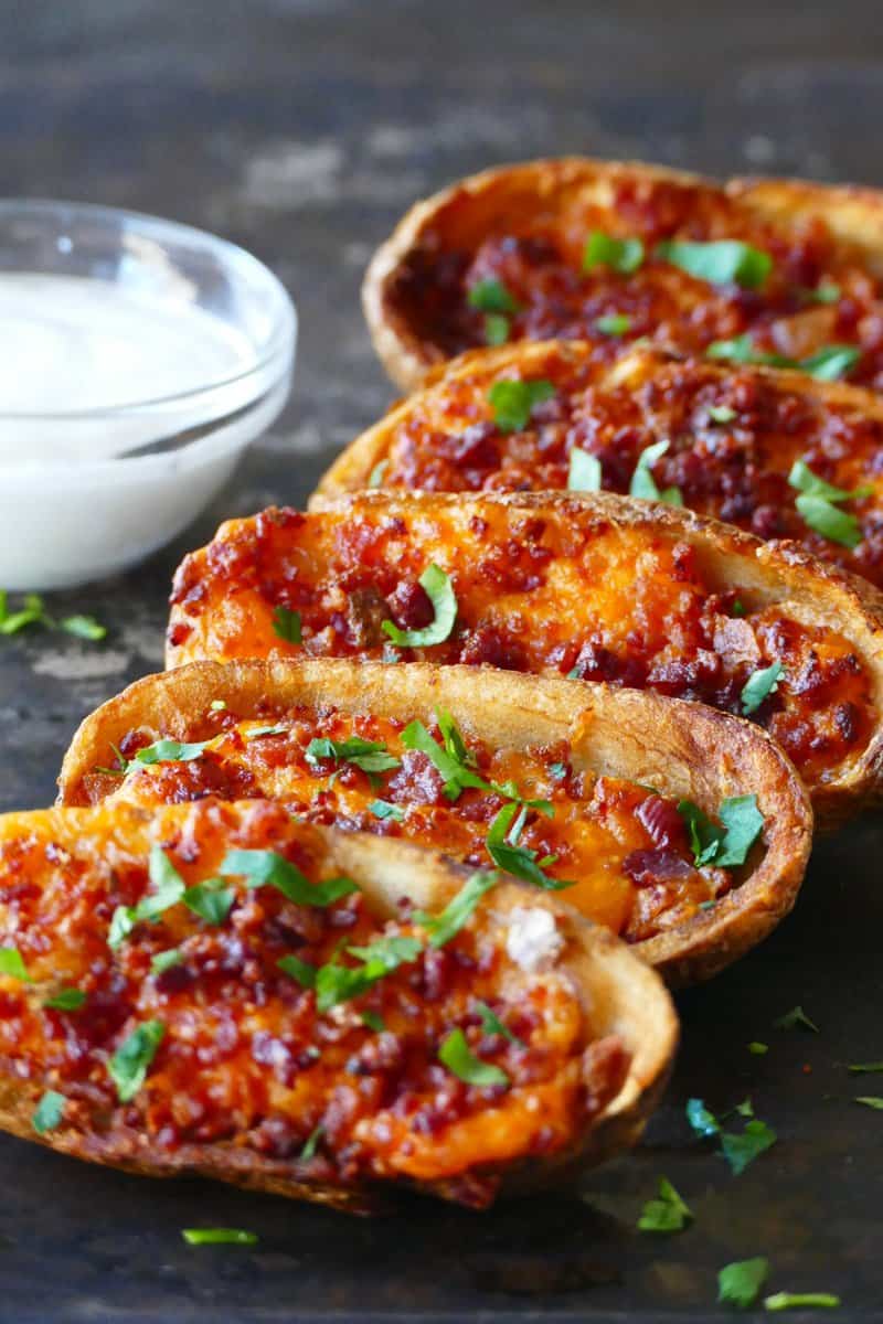 Air fryer frozen potato skins in a row with melted cheese and bacon and garnished with parsley with a glass bowl of white sauce.