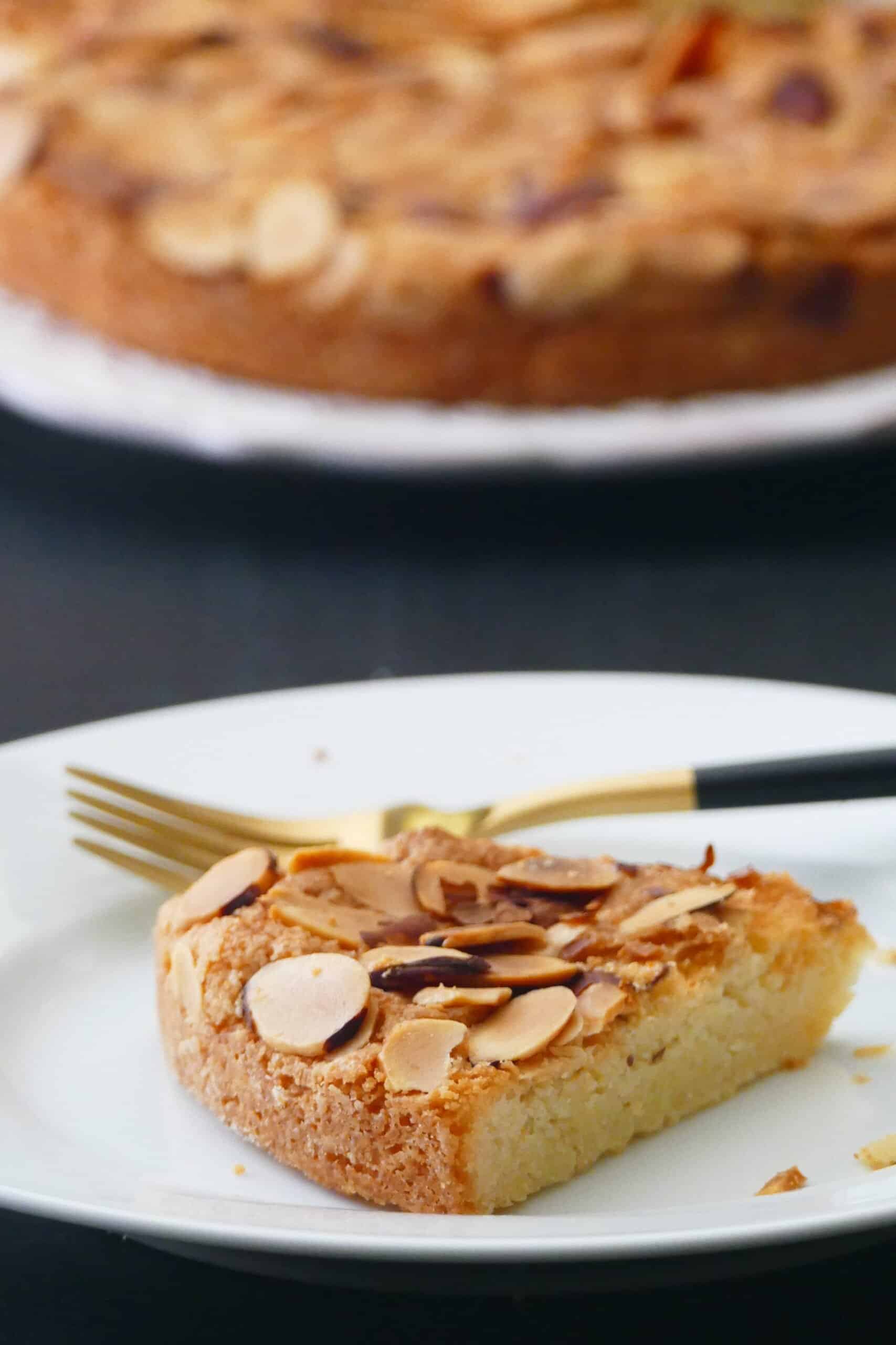 Air fryer almond cake - Slice of cake on white plate with slivered almonds baked to a golden brown and whole cake in back ground