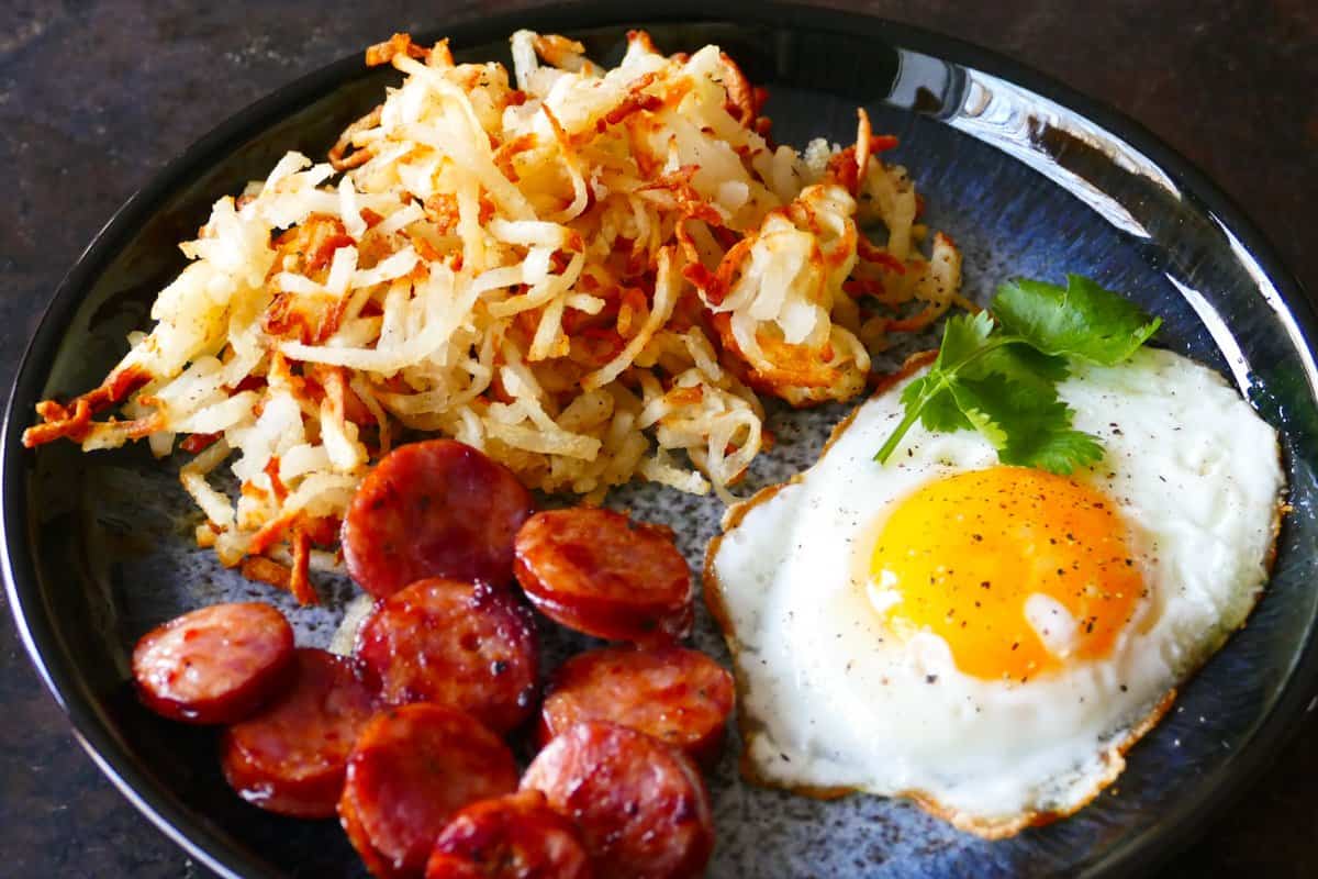 Blue plate with golden brown shredded hashbrowns, fried egg and sliced sausage.