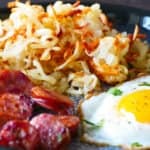 Blue plate with golden brown shredded hashbrowns, fried egg and sliced sausage.