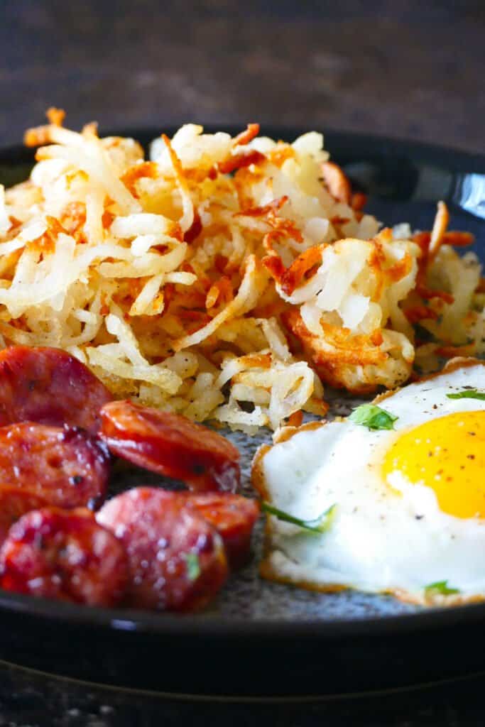 Blue plate with golden brown shredded hashbrowns, fried egg and sliced sausage.