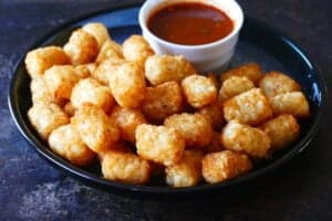 Air fryer tater tots stacked on a dark plate with a bowl of red sauce