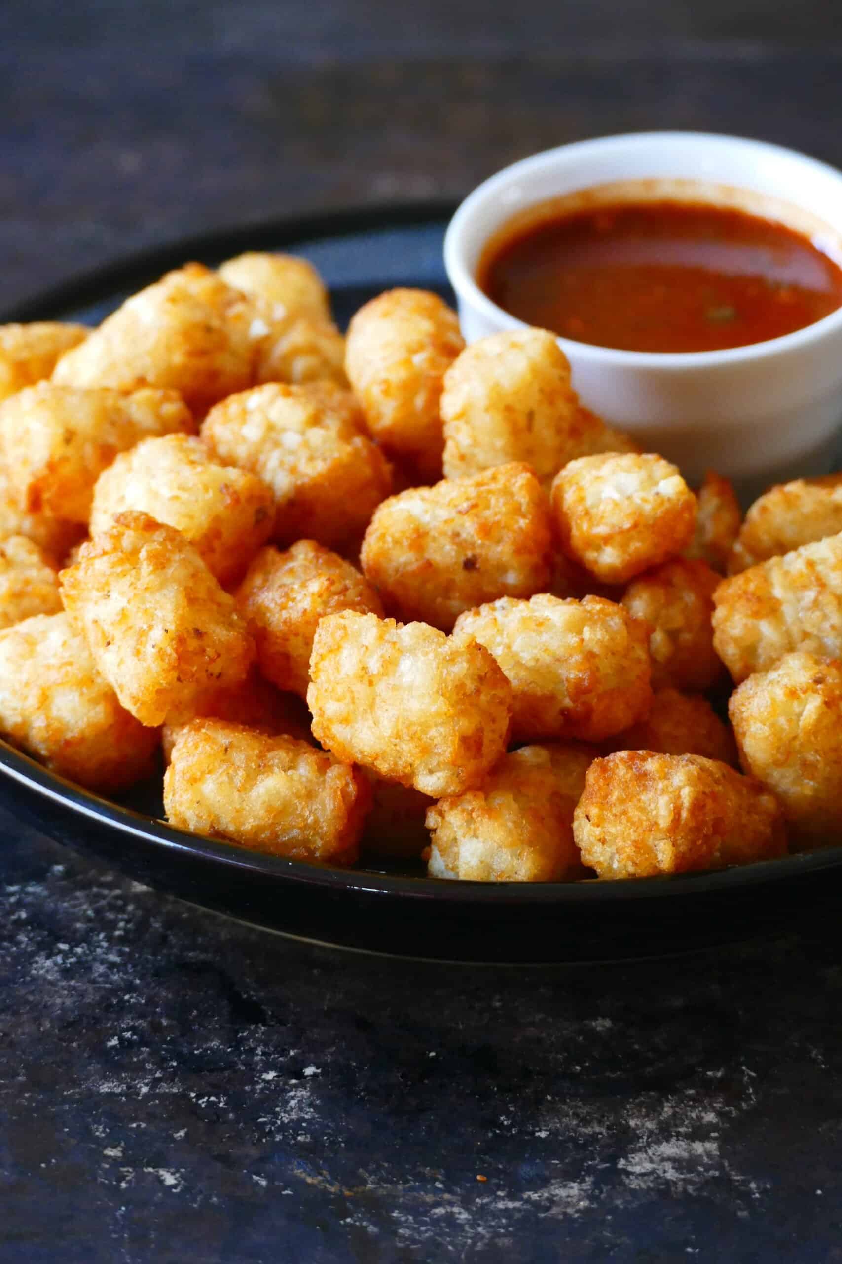 Air fryer tater tots stacked on a dark plate with a bowl of red sauce
