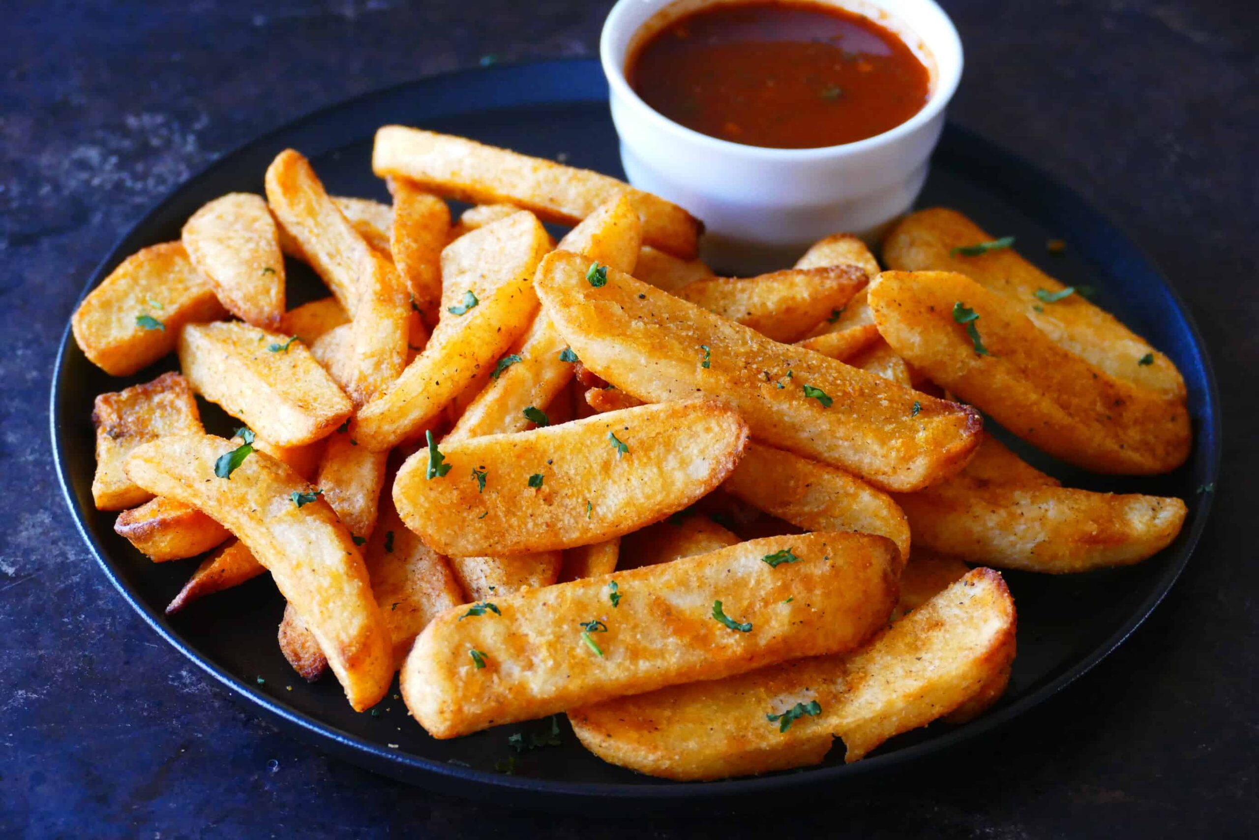 Black plate with steak fries garnished with parsley and bowl of red sauce