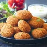 Golden brown, crunchy falafel stacked on a plate with lettuce tomato, white sauce and lemon in background