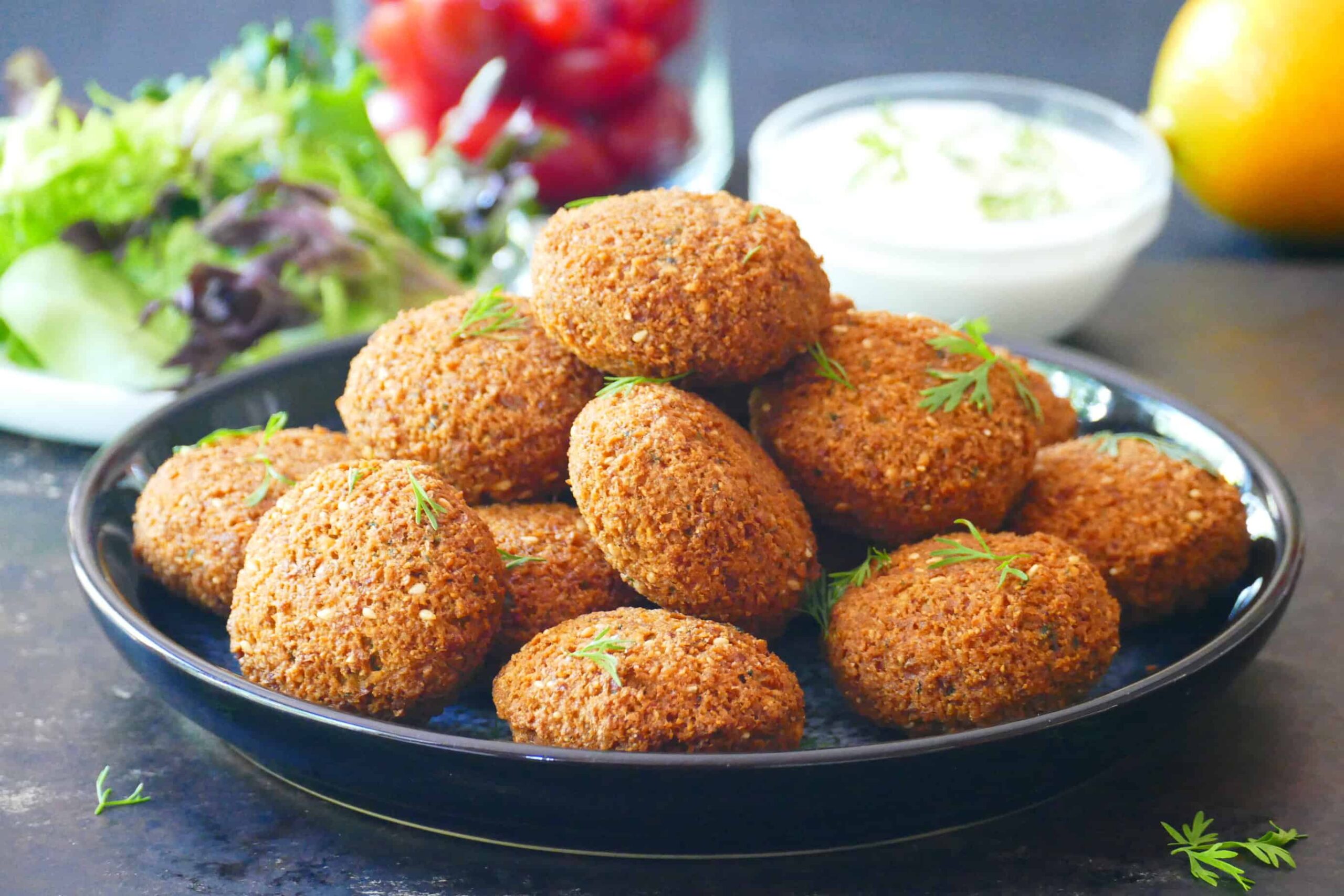 Golden brown, crunchy falafel stacked on a plate with lettuce tomato, white sauce and lemon in background