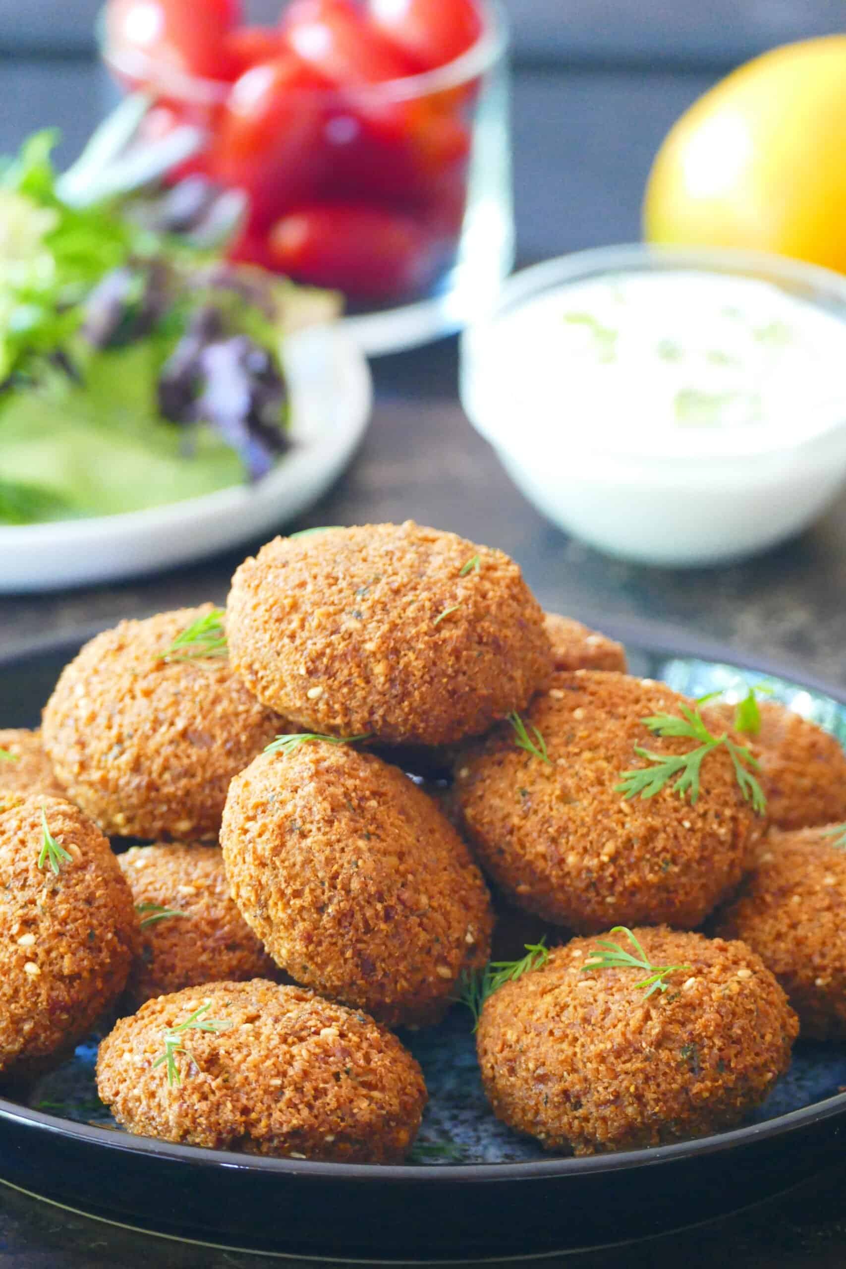 Golden brown, crunchy falafel stacked on a plate with lettuce tomato, white sauce and lemon in background
