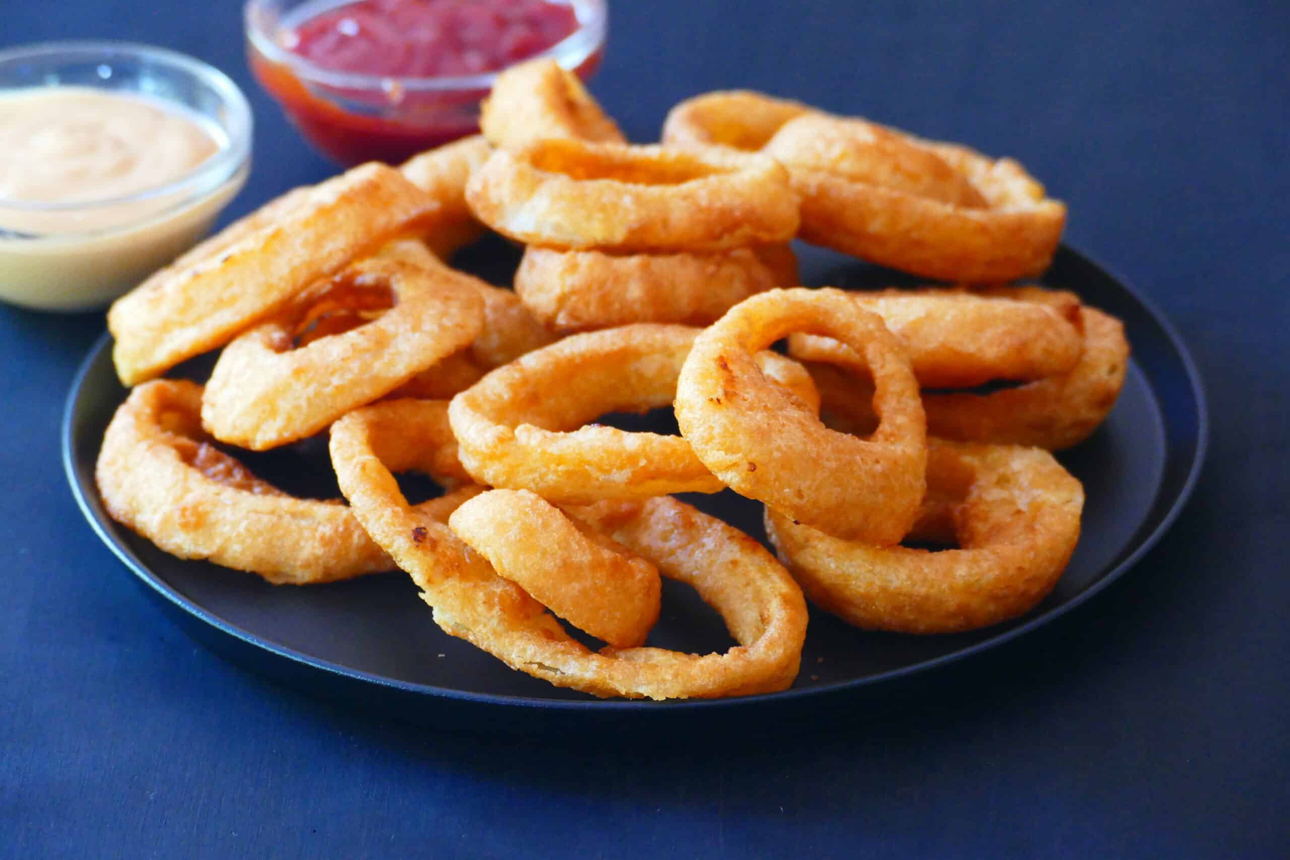 Onion rings on a black plate with red and white sauces in background