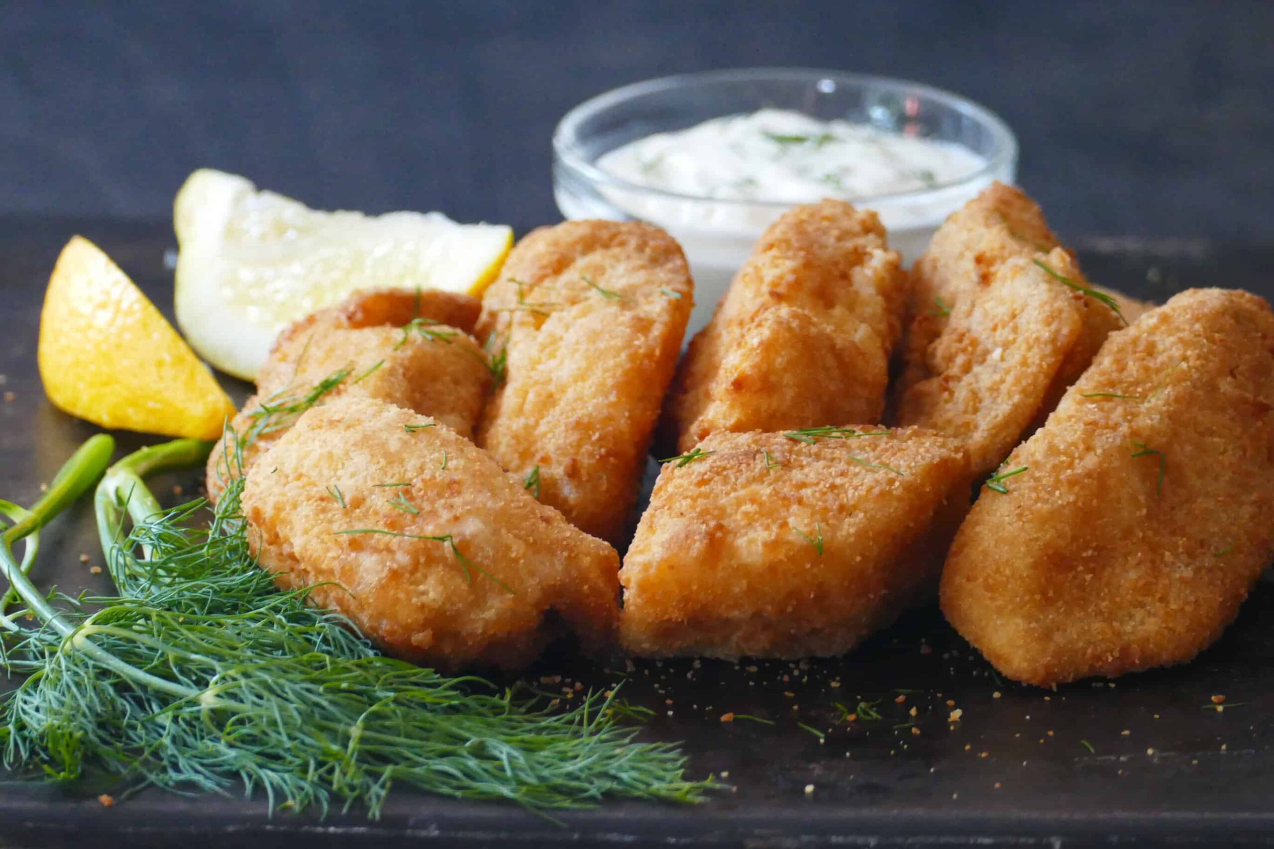 Golden fish fillets on dark surface with white sauce, dill and lemon