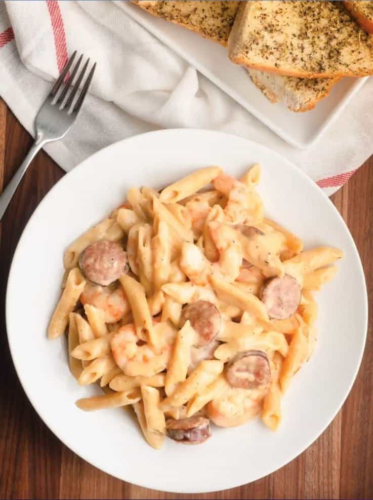 Tubular pasta with sausage and shrimp on a white plate with garlic bread in background