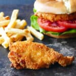 A piece of breaded chicken with french fries and a chicken sandwich in the background.