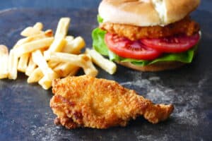 A piece of breaded chicken with french fries and a chicken sandwich in the background.