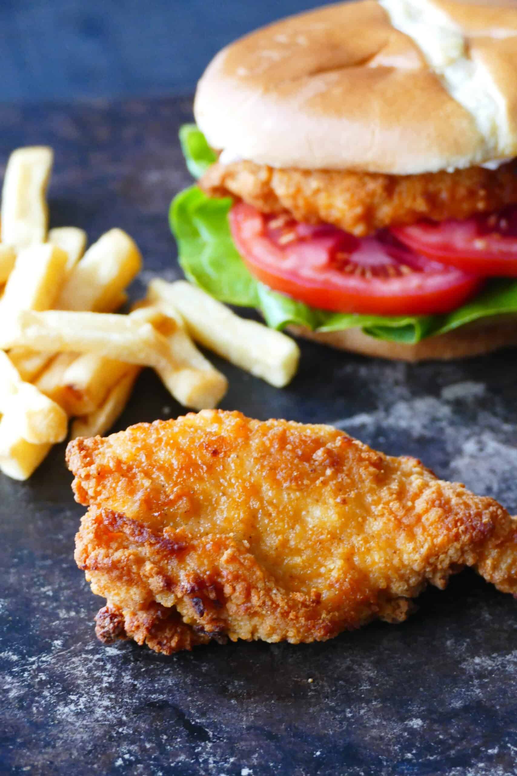 Air fryer frozen breaded chicken breast - A piece of breaded chicken with french fries and a chicken sandwich in the background.