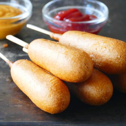 Five stacked cooked corn dogs with ketchup and mustard in bowls.