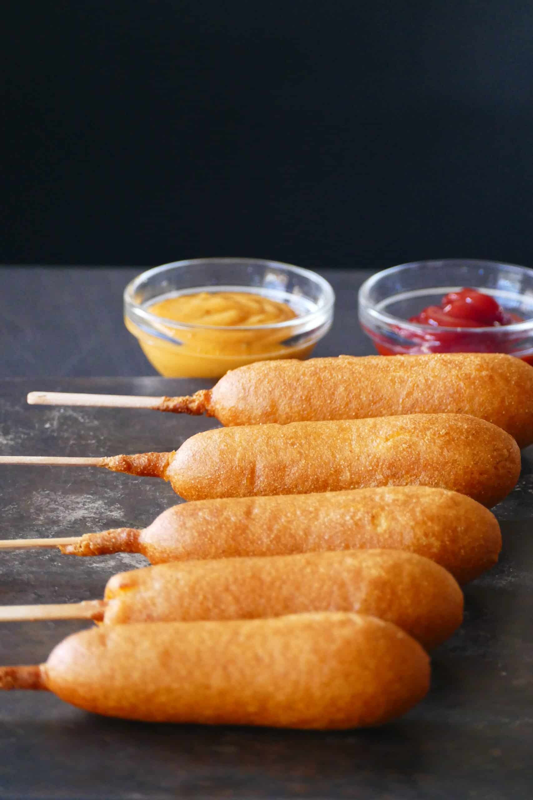 Air fryer frozen corn dogs - Row of five cooked corn dogs with ketchup and mustard in bowls.