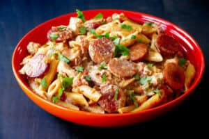 Red bowl with pasta, sausage, chicken, garnished with parsley.