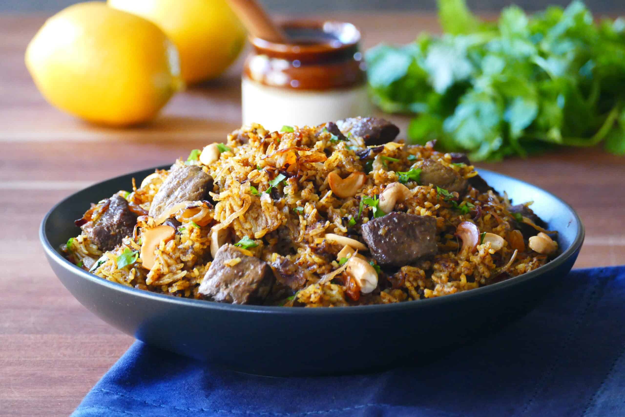 Instant Pot lamb biryani in a black bowl has rice, cashews, pieces of browned lamb on a blue napkin.