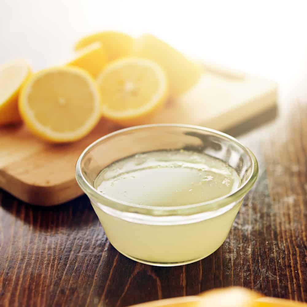 Lemon juice in glass bowl with cut lemons in background.