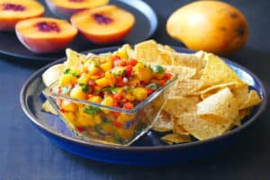 Mango peach salsa in glass bowl with bell peppers, cilantro, red onions and tortilla chips, peach and mango in background.