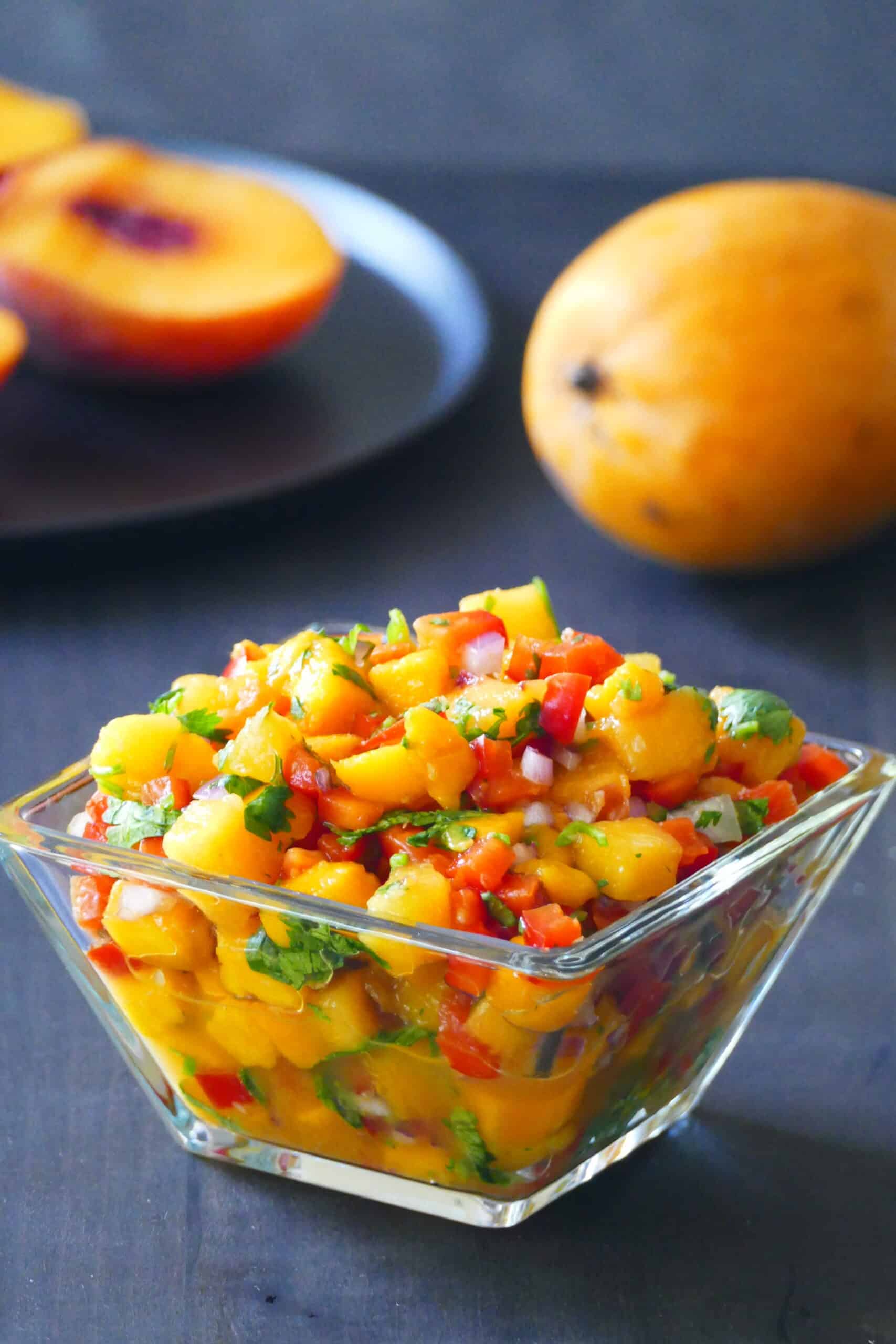 Mango peach salsa in glass bowl with bell peppers, cilantro, red onions and peach and mango in background.