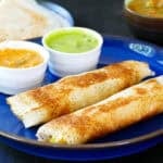 Two masala dosas on plate and green and red chutneys and dosas and sambar in background.